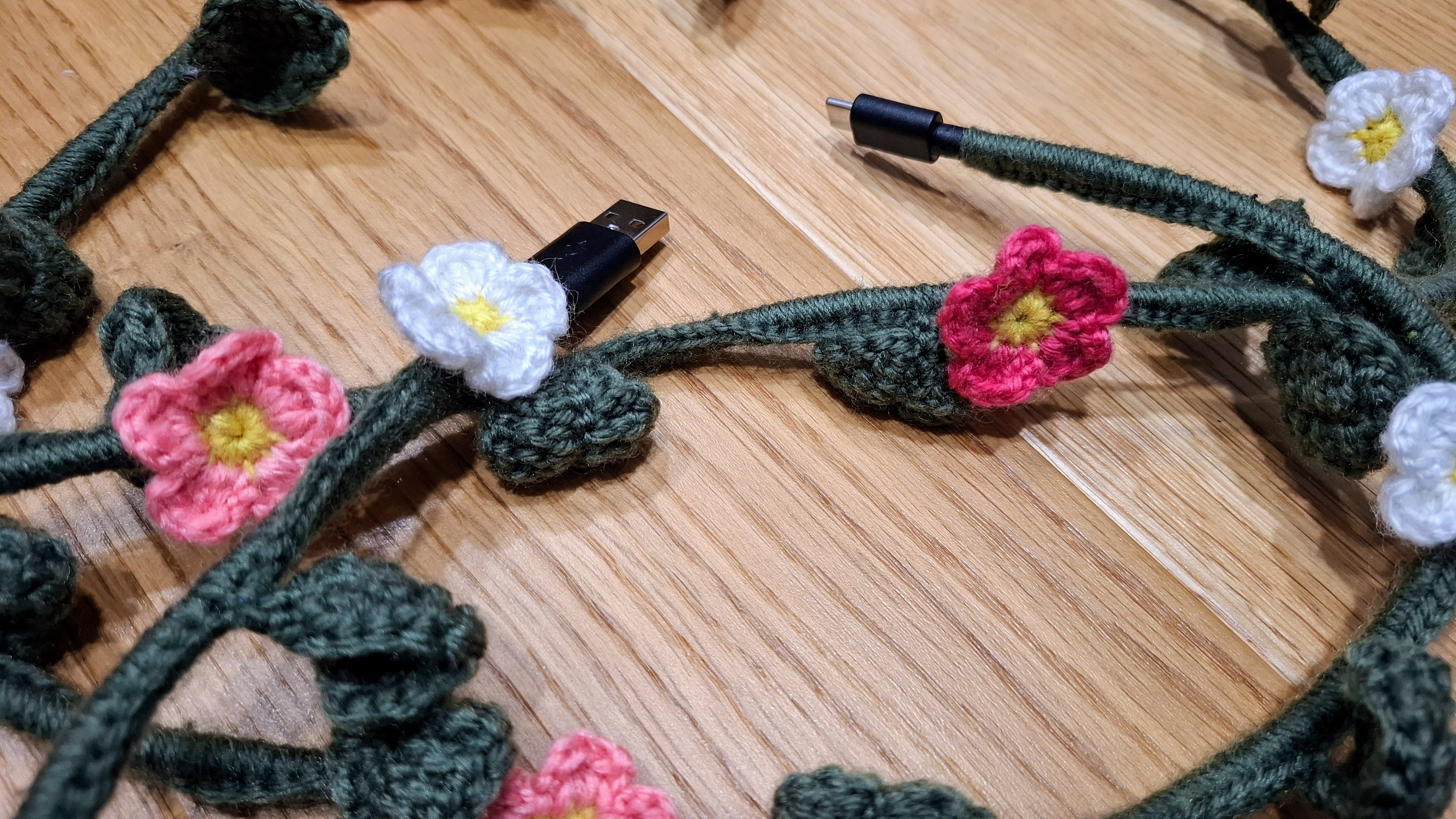 A crocheted floral vine wrapped around a charging cable, with pink and white flowers